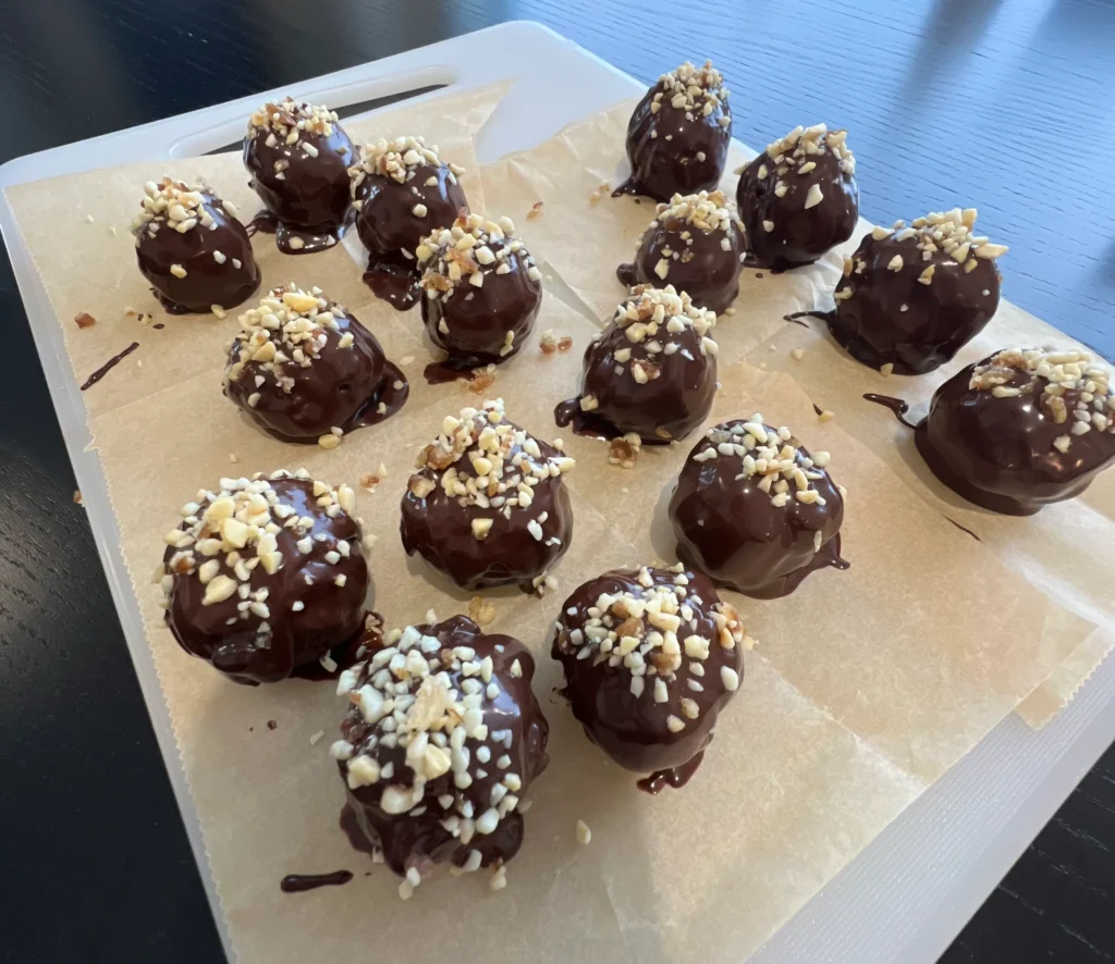 Energy balls dipped in melted chocolate and garnished with crumbled nuts, on a cutting board.
